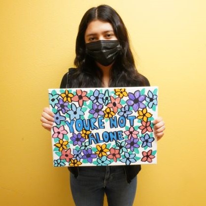 Rubi, youth, holds colorful sign with hand-drawn flowers and statement: You're Not Alone.